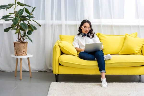 Thoughtful asian freelancer in glasses using laptop while sitting comfortably on yellow sofa at home — Stock Photo
