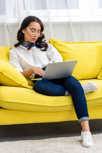 Selective focus of concentrated asian freelancer in glasses using laptop while sitting on yellow sofa at home — Stock Photo