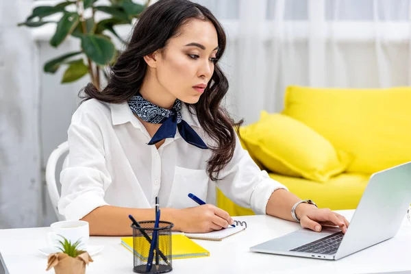 Selective focus of attentive asian freelancer using laptop and writing in notebook — Stock Photo