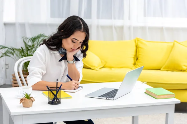 Young asian freelancer using laptop and writing in notebook, supporting head with hand — Stock Photo