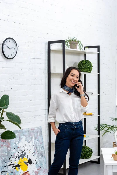 Lächelnder asiatischer Freiberufler, der mit dem Smartphone spricht und die Hand in der Tasche hält, während er zu Hause vor dem Regal steht — Stockfoto