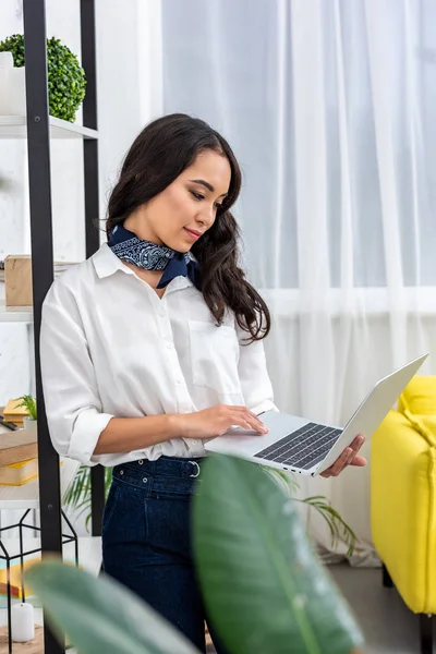 Belle asiatique pigiste en utilisant ordinateur portable tout en se tenant près de lieu de travail à la maison — Photo de stock