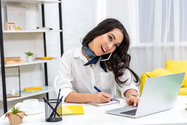 Smiling asian freelancer using laptop and talking on smartphone while writing in notebook — Stock Photo