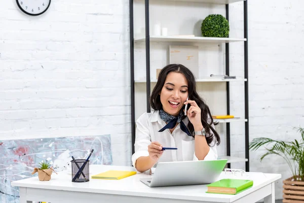 Smiling asian freelancer talking on smartphone and holding pen while sitting at home workplace — Stock Photo
