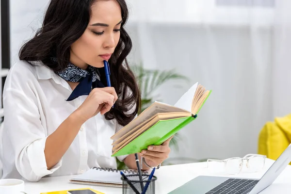 Selective focus of concentrated asian freelancer reading book and holding pen near lips while sitting at work table — Stock Photo