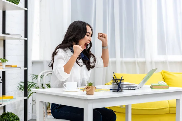 Exhausted asian freelance yawning and raising hands while sitting at work desk at home — Stock Photo