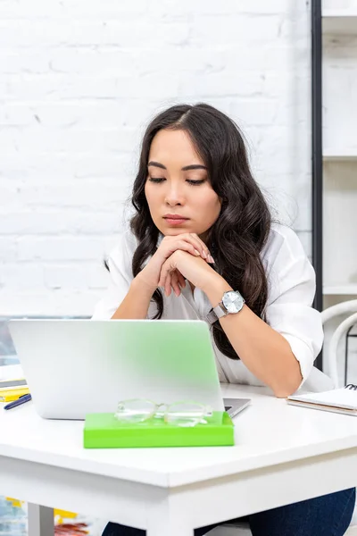 Tired asian freelancer looking at laptop screen and clutching hands under chin — Stock Photo