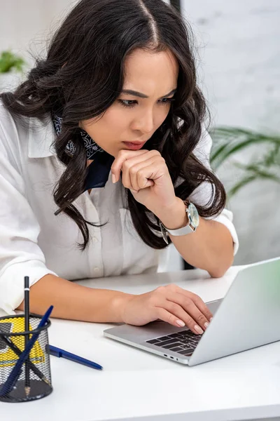 Serious freelancer attentively using laptop at home workplace and holding hand under chin — Stock Photo