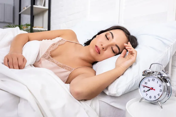 Hermosa mujer joven en camisón elegante durmiendo en ropa de cama blanca, mientras que el reloj despertador que muestra cuarto a las ocho de la mañana — Stock Photo