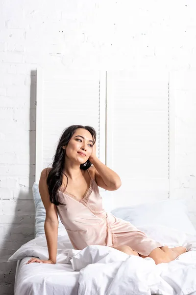 Attraktive junge asiatische Frau im eleganten Nachthemd sitzt auf Bettwäsche und hält Hand auf Kopf — Stockfoto