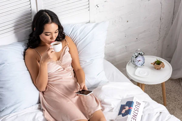 Молодая азиатка в элегантной ночной рубашке пьет кофе и пользуется смартфоном во время пребывания в постели — стоковое фото