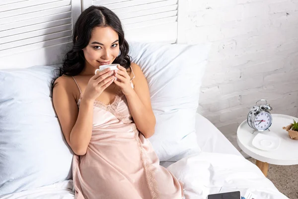 Beautifil mulher asiática em elegante nighty beber café enquanto ficar na cama e olhando para a câmera — Fotografia de Stock