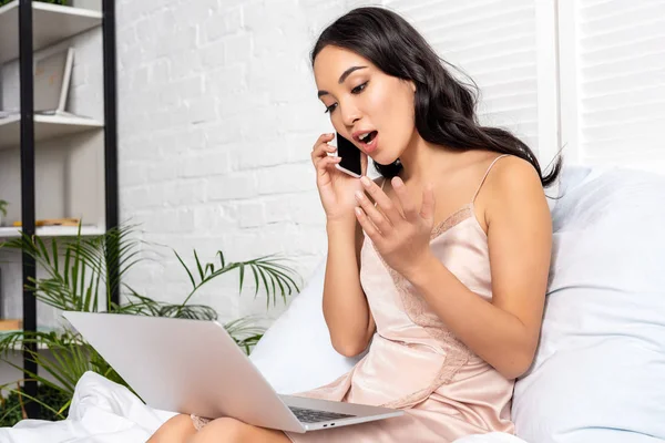 Hermosa asiática freelancer utilizando el ordenador portátil y emocionalmente hablando en el teléfono inteligente durante su estancia en la cama - foto de stock