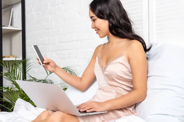 Bella asiatica freelance utilizzando il computer portatile e tenendo smartphone durante il soggiorno a letto — Foto stock