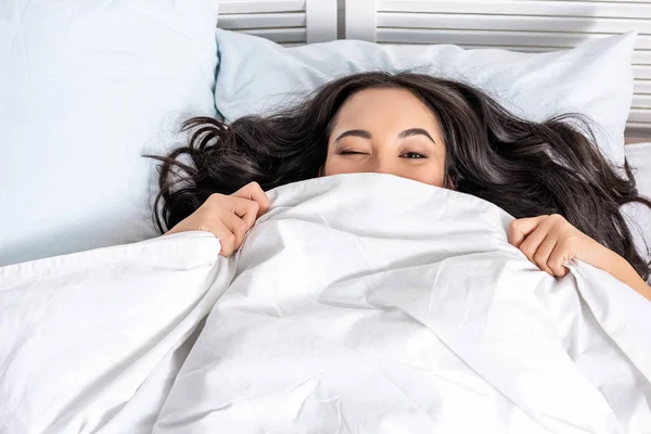Young beautiful asian woman winking while looking out of blanket — Stock Photo