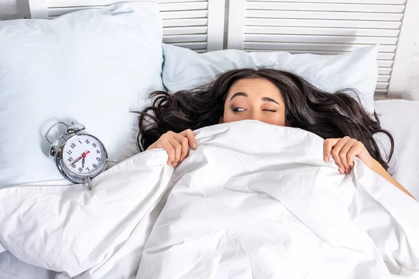 Young beautiful asian woman looking at alarm clock from under the blanket with squinting one eye — Stock Photo