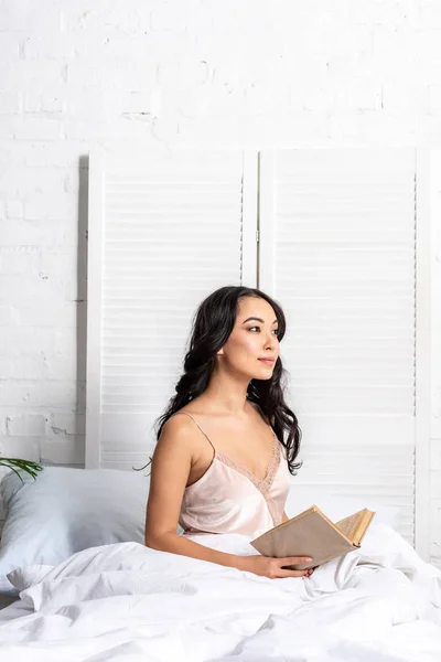 Bastante asiático mujer en elegante camisón sentado en cama y holding libro - foto de stock