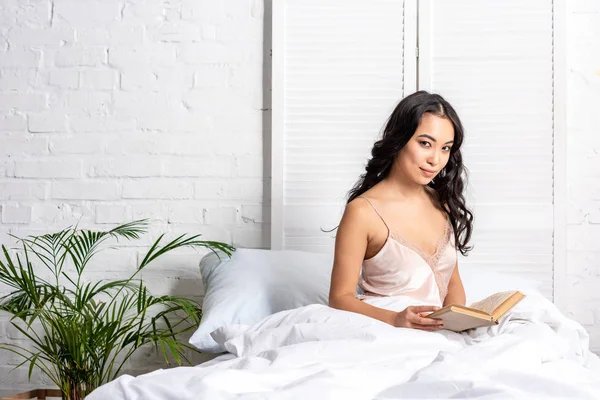 Hübsche asiatische Frau im eleganten Nachthemd sitzt im Bett und hält Buch, während sie in die Kamera schaut — Stockfoto