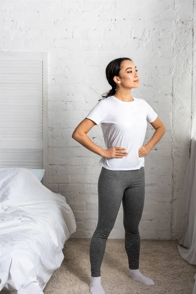 Jovem mulher asiática em t-shirt branca e leggings cinza de pé no quarto segurando as mãos para quadris — Fotografia de Stock