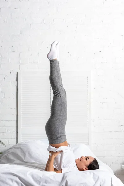 Ásia mulher no branco t-shirt e cinza leggings fazendo ombro stand pose exercício no cama — Fotografia de Stock