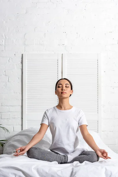 Asian woman in white t-shirt and grey leggings sitting in lotus pose in bed — Stock Photo