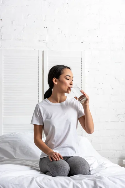 Atraente asiático menina no branco t-shirt sentado no cama e beber água — Fotografia de Stock