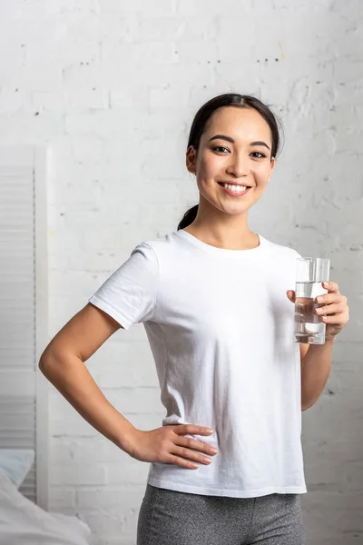 Hermosa joven mujer asiática con vaso de agua de pie en el dormitorio con la mano en la cadera y mirando a la cámara - foto de stock