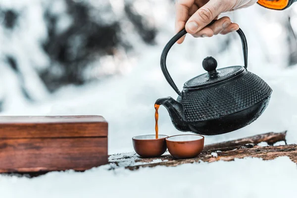 Cropped view of male traveler pouring tea from kettle in cups in winter forest — Stock Photo