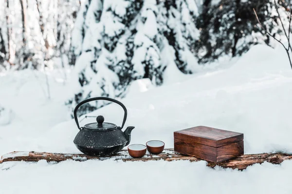 Kettle with small cups on wooden surface in snowy forest — Stock Photo