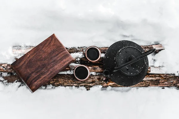 Вид сверху на чайник, чашки с чаем и деревянный ящик на бревнах среди снега — стоковое фото