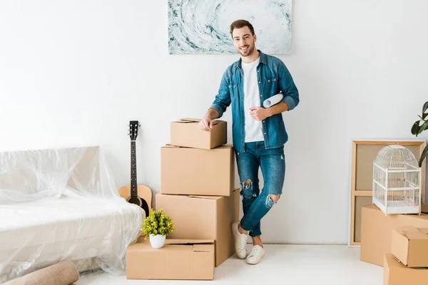 Full length view of smiling man in jeans standing near cardboard boxes at new home — Stock Photo