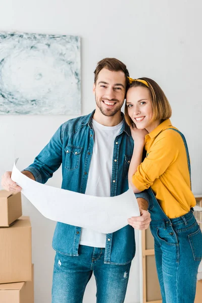 Lachendes Paar in Jeans mit Blaupause und Blick in die Kamera zu Hause — Stockfoto