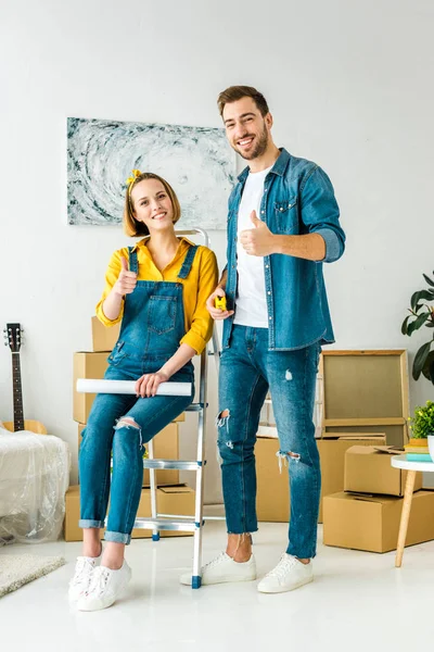 Visão comprimento total do casal com caixas de papelão e planta mostrando polegares para cima — Fotografia de Stock