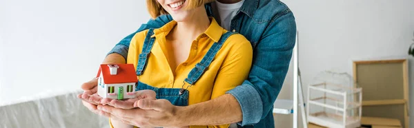 Panoramic shot of smiling couple holding toy house and looking at camera at home — Stock Photo