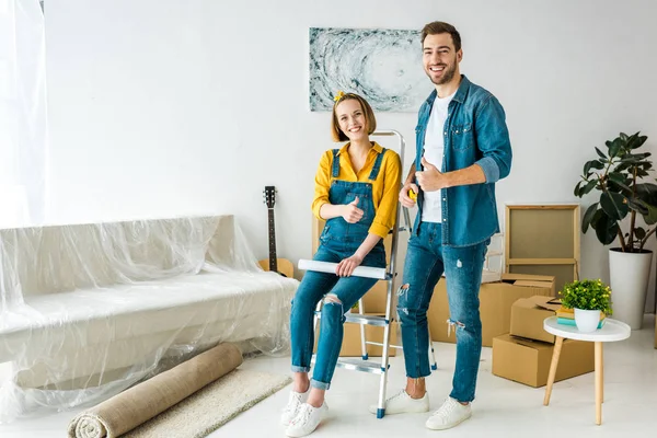 Full length view of couple with cardboard boxes and blueprint showing thumbs up — Stock Photo