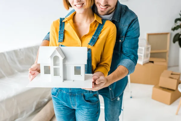 Cropped view of smiling couple holding toy house and looking at camera at home — Stock Photo