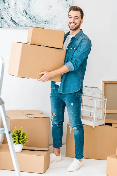 Vista completa del hombre riendo en jeans sosteniendo cajas de cartón y mirando a la cámara - foto de stock