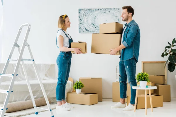 Full length view of smiling couple holding cardboard boxes and looking at each other at home — Stock Photo