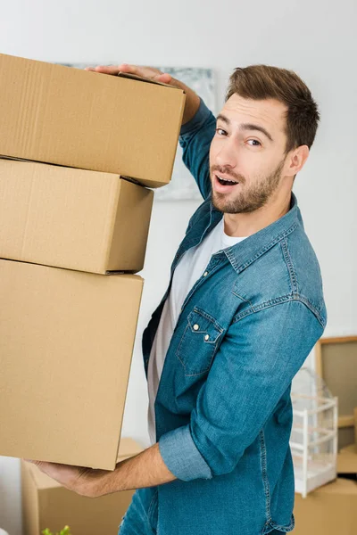 Hombre alegre en chaqueta de mezclilla sosteniendo cajas de cartón y mirando a la cámara - foto de stock