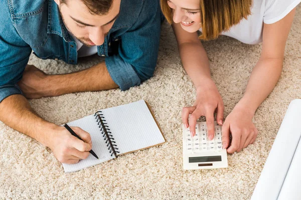 Vista cortada de casal alegre deitado no tapete com caneta, caderno e calculadora — Fotografia de Stock