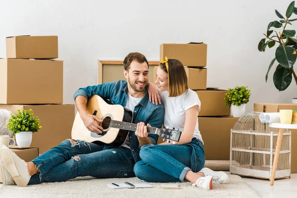 Sorrindo homem sentado no tapete com a esposa e tocando guitarra acústica — Fotografia de Stock