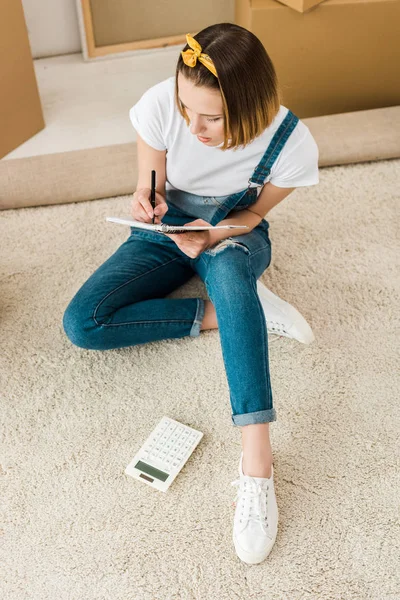 Overhead view of concentrated girl writing in notebook while sitting on carpet — Stock Photo