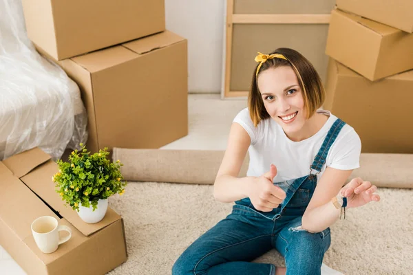 Hochwinkelaufnahme einer Frau, die Schlüssel in der Hand hält und den Daumen in der Nähe von Kartons zeigt — Stockfoto