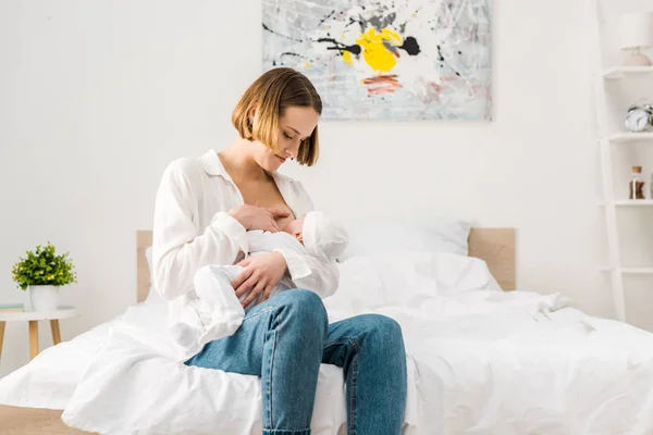 Мама в джинсах сидит на кровати и кормит грудью ребенка — стоковое фото