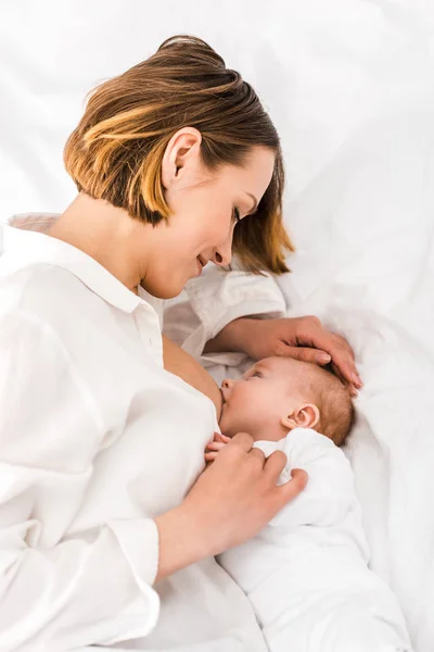 Мать в белой рубашке лежит на кровати и кормит грудью ребенка — стоковое фото