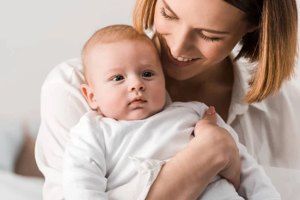 Sonriente joven madre en camisa blanca sosteniendo al bebé en casa - foto de stock