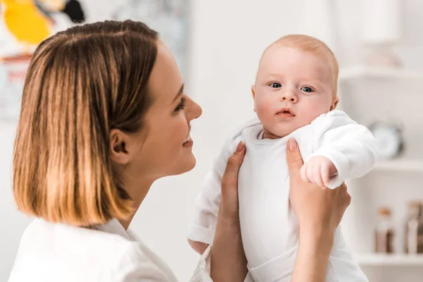 Sorrindo jovem mãe em camisa branca segurando bebê em casa — Fotografia de Stock