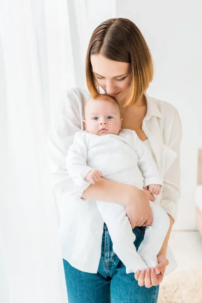 Sonriente joven madre en camisa blanca sosteniendo al bebé en casa - foto de stock