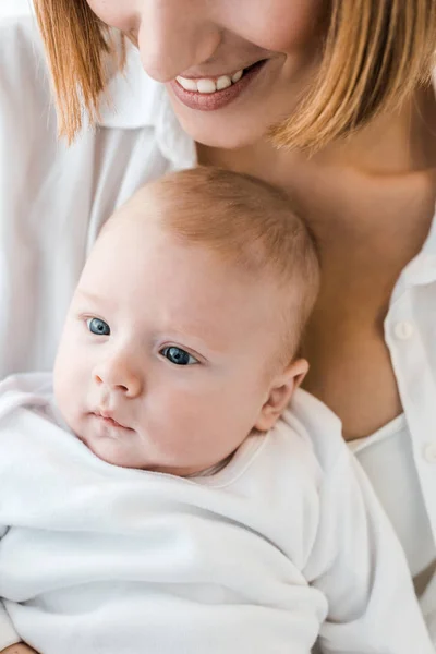 Vista ritagliata della madre sorridente in camicia bianca che tiene il bambino a casa — Foto stock