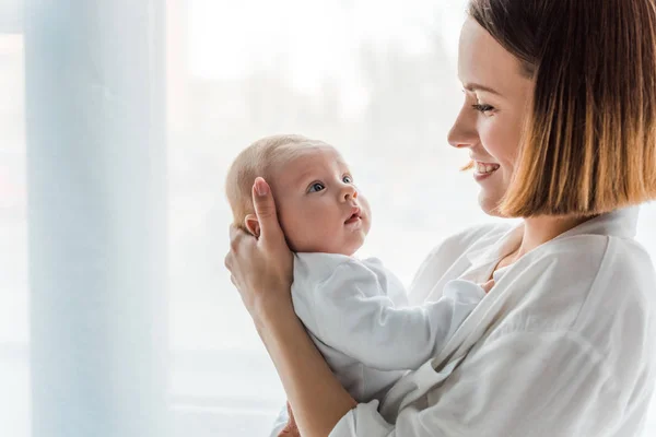 Mãe sorridente em camisa branca segurando bebê em casa — Fotografia de Stock